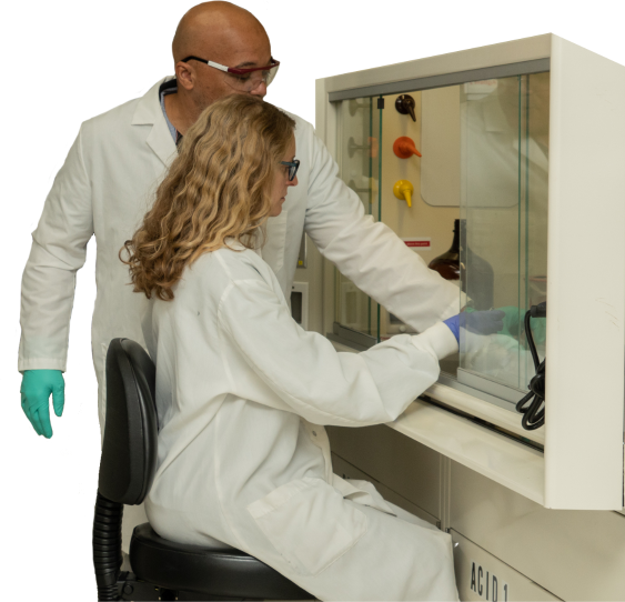 Two people in lab coats and gloves working at a fume hood